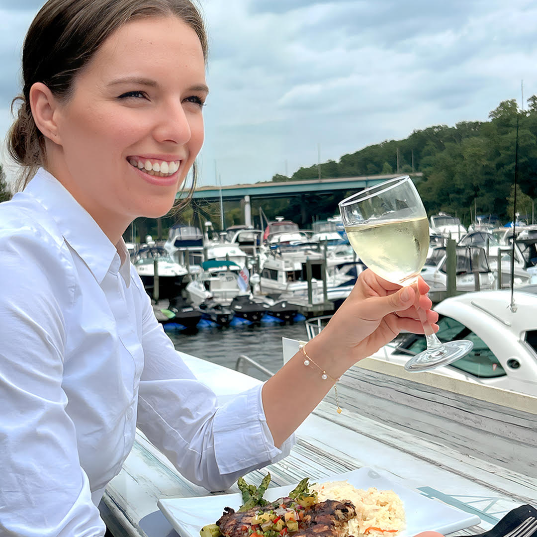 A female customer of The Harbour Grille, a restaurant in Woodbridge, VA, enjoying a glass of wine in her hand, smiling, with a beautiful background of parked boats. 